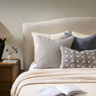 A styled bed with a beige upholstered headboard, cream bedding, and multiple decorative pillows including striped, navy floral, and solid navy options. A wooden bedside table with a lamp and small vase sits to the left, and an open book rests on the blanket in the foreground.